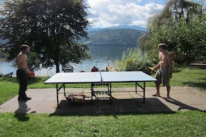 Twee personen spelen tafeltennis buiten, met een meer en bergen op de achtergrond.