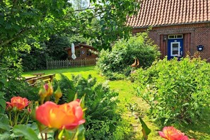 A brick building with a blue door, surrounded by lush greenery and a wooden fence. - Klaus-Störtebeker-House (Neuharlingersiel)