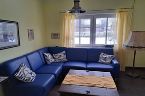 A room with a blue leather sofa, a wooden coffee table, a window with curtains, and a lamp. - Klaus-Störtebeker-House (Neuharlingersiel)