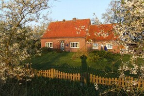 Une maison en briques avec un toit rouge, entourée d’une clôture en bois et bordée d’arbres en fleurs.
