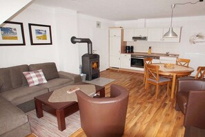A living room with a sofa, coffee table, and a fireplace. There is a kitchen area with cabinets and a dining table. - 07 Sommerhus Fredericia (Burg auf Fehmarn)