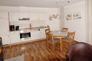 A compact kitchen with white cabinets, a built-in oven, and a dining area with a round table and four chairs. - 07 Sommerhus Fredericia (Burg auf Fehmarn)
