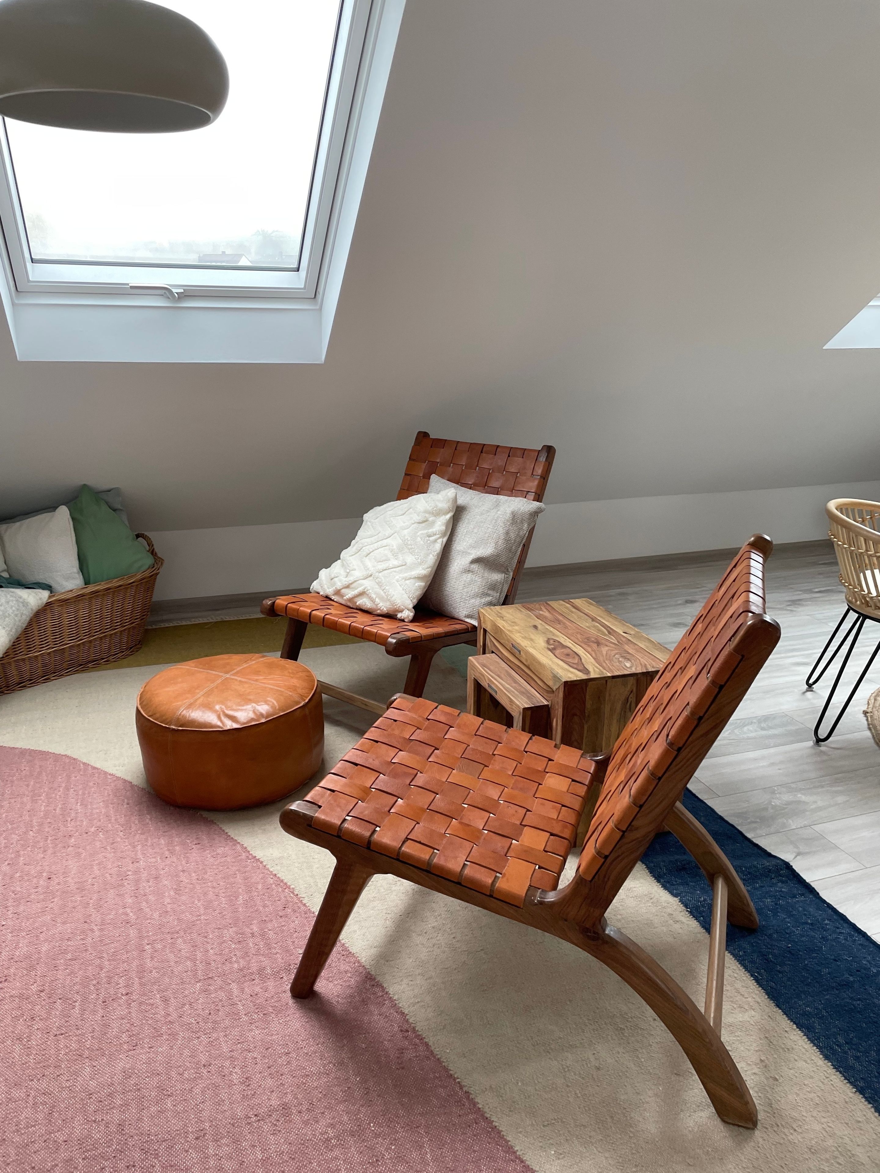 Une pièce avec un plafond haut, une chaise en bois avec un dossier en osier, une petite table en bois, un pouf rond en cuir et un panier en osier.