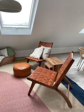 A room with a high ceiling, a wooden chair with a woven backrest, a small wooden table, a round leather ottoman, and a wicker basket.