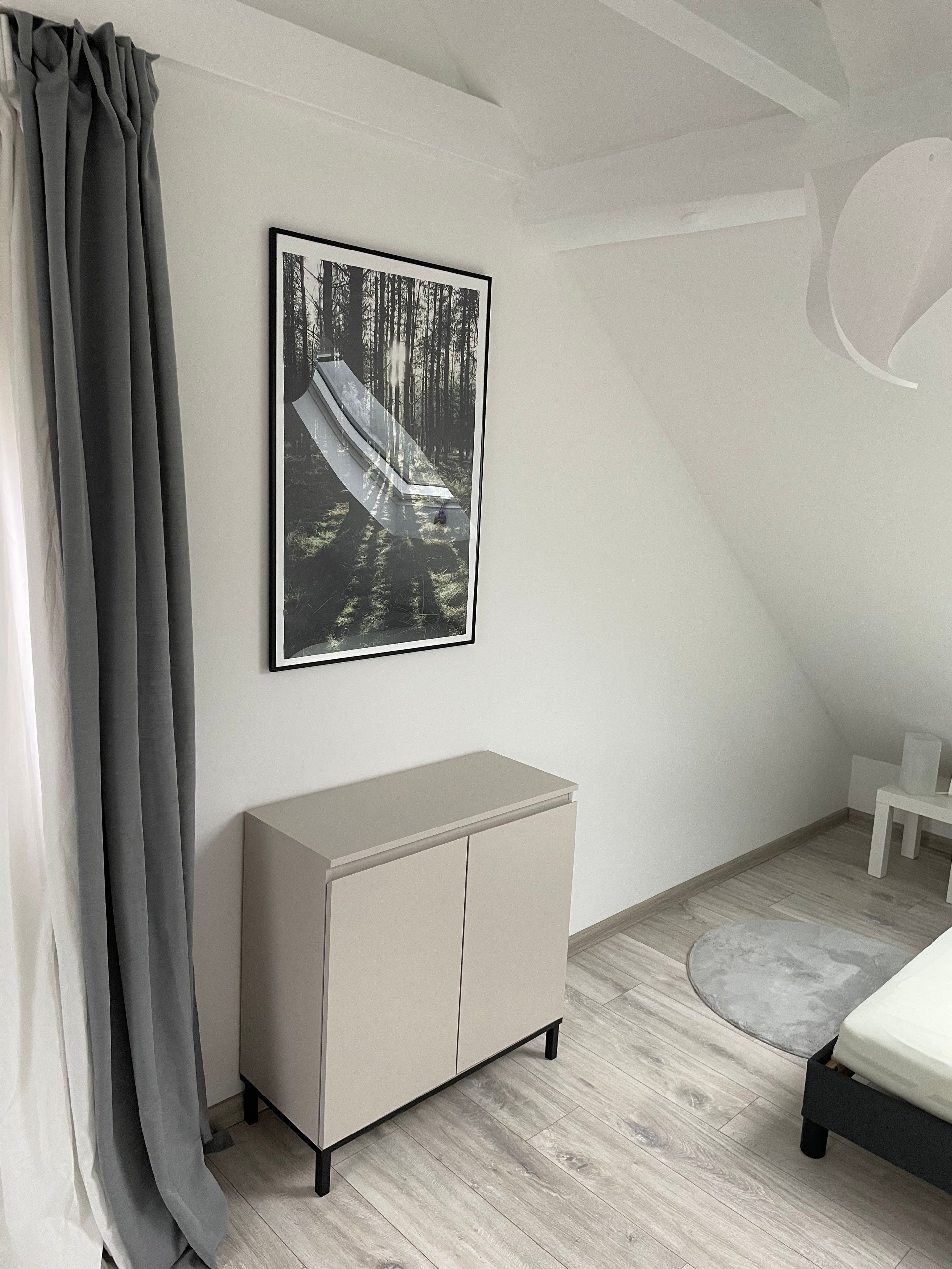 Une chambre à coucher avec un lit, une armoire grise, un tableau encadré représentant une forêt et un rideau gris.