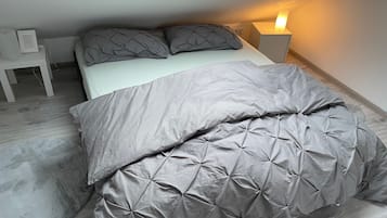 Ein ordentlich bezogenes Bett mit einer grauen Bettdecke und Kissen, ein Nachttisch mit einer Lampe und ein kleiner weißer Stuhl.