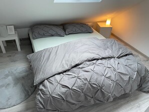 A neatly made bed with a grey duvet and pillows, a bedside table with a lamp, and a small white chair.