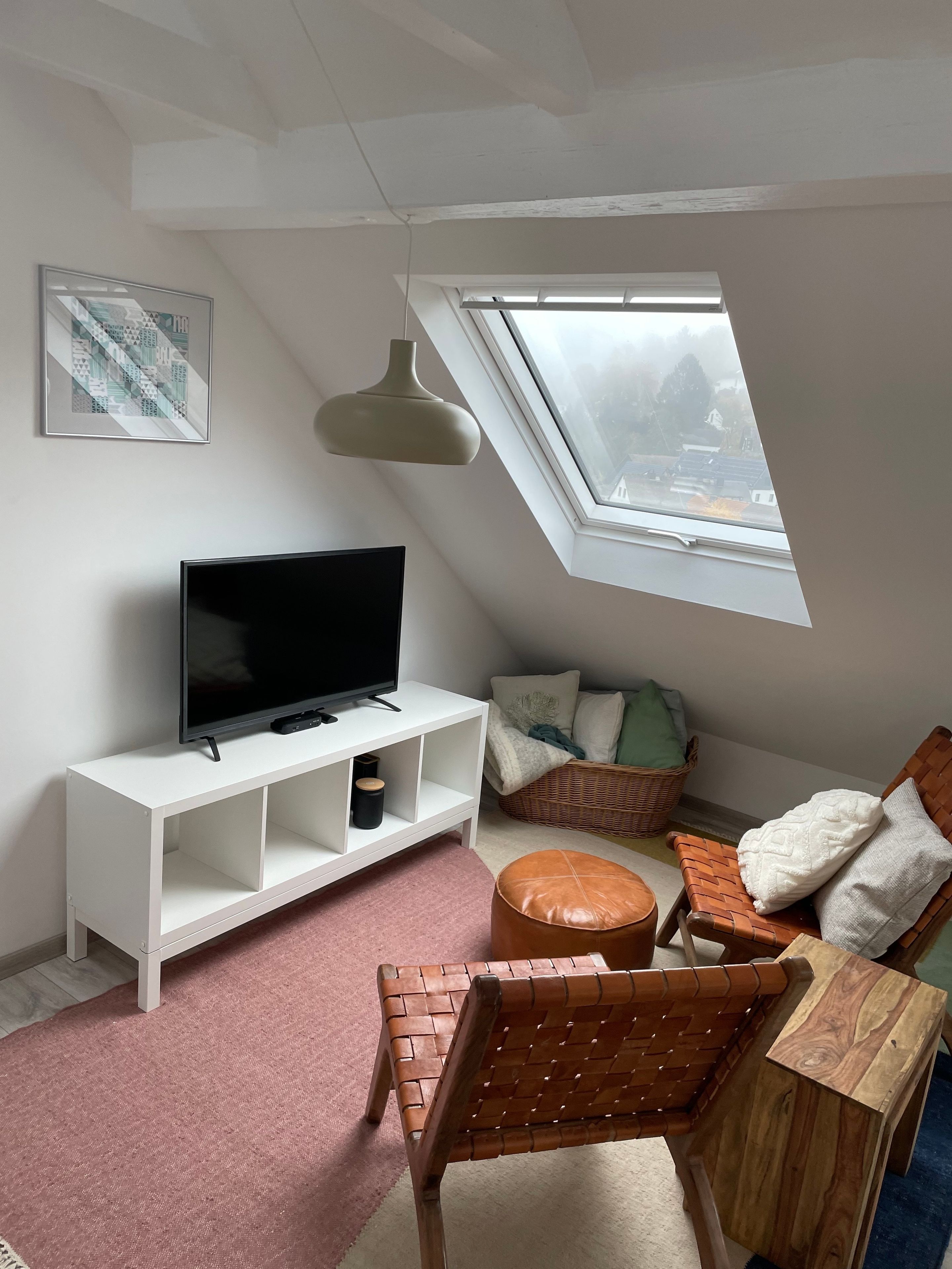 Ein Loft-Zimmer mit schrägem Dach, einem Fernseher auf einer weißen Konsole, einem Korbstuhl, einem Holztisch und Blick durch ein Oberlicht.