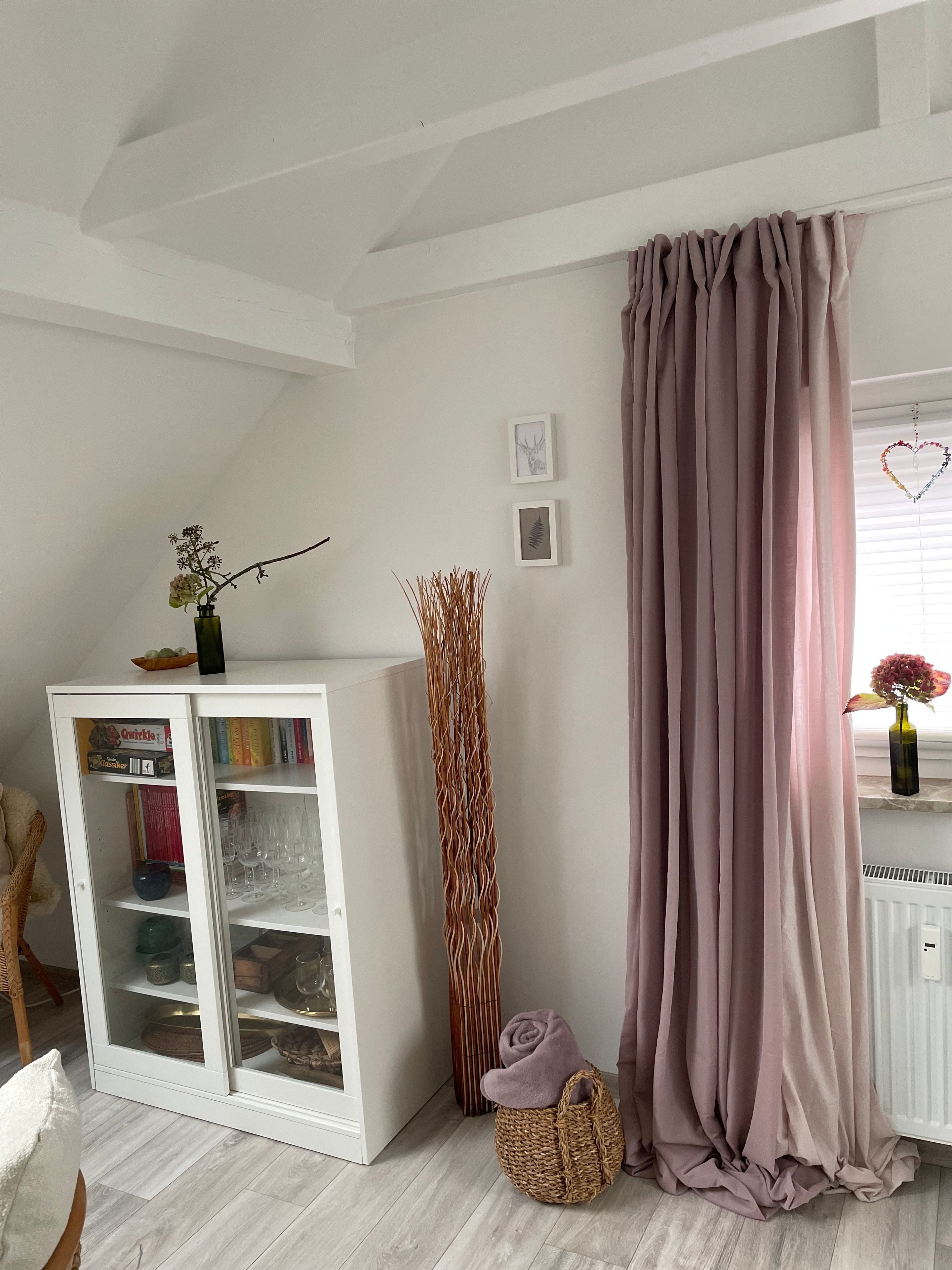 A room with a white cabinet, a tall decorative plant, a pink curtain, and a small basket with a rolled towel.