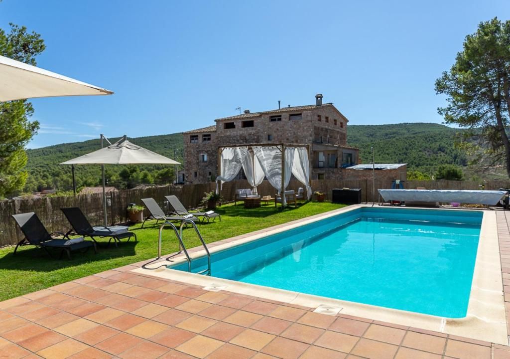 A swimming pool with lounge chairs and umbrellas, a stone building in the background, and a hilly landscape.