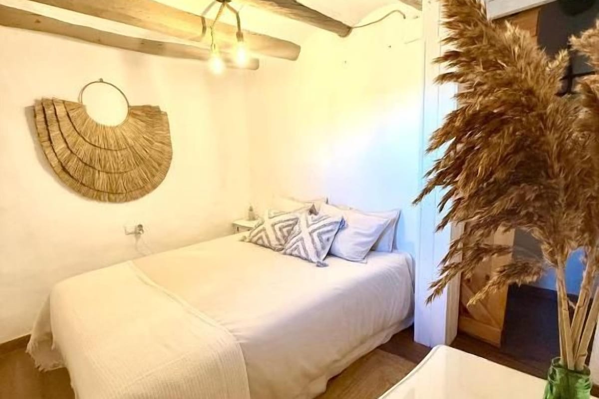A bedroom with a single bed, a bedside table, a green vase with dried plants, and a wall-mounted light fixture.