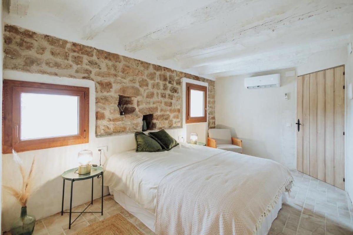 A bedroom with a stone wall, a bed with white bedding, a bedside table with a lamp, and a wooden door.