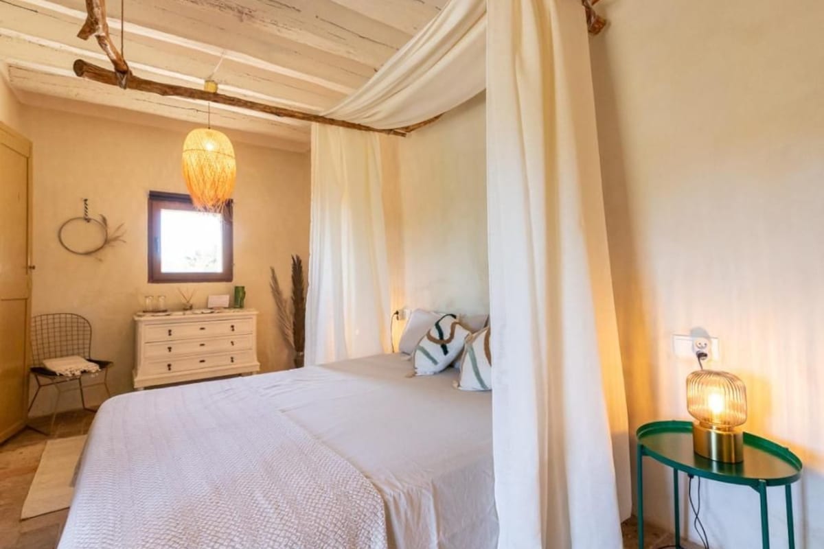 A bedroom with a bed, a dresser, a chair, a bedside table with a lamp, and a window with curtains.