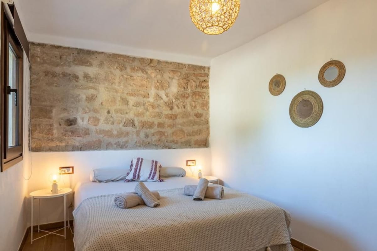 A bedroom with a stone wall, a bed with rolled towels, bedside lamps, and wall-mounted decor.
