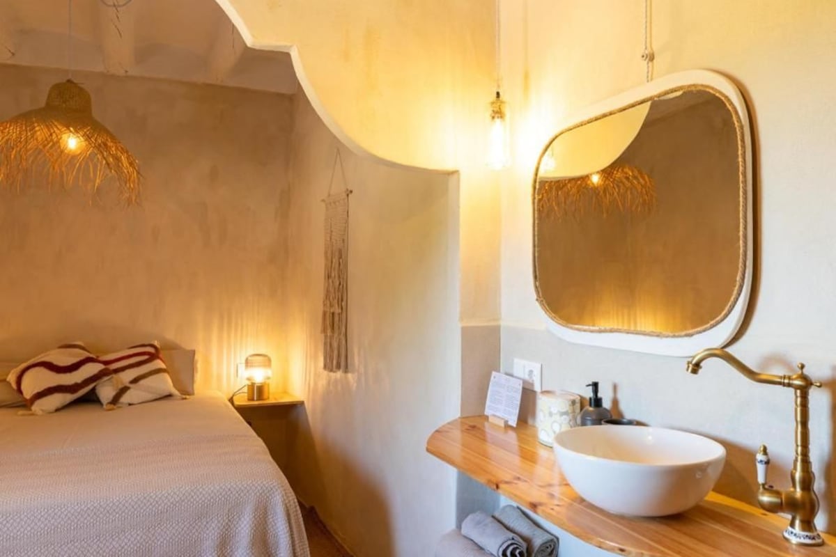 A hotel room with a bed, bedside lamp, wooden nightstand, and a large mirror with a brass faucet.
