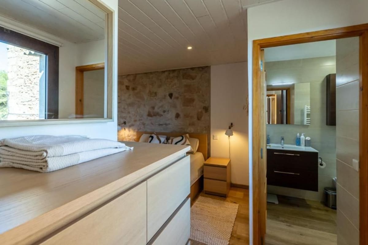 A bedroom with a wooden headboard, a bedside table, a lamp, and a view of the bathroom through an open door.