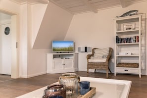 A room with a television on a stand, a wicker chair, a bookshelf, and a door with a circular window. - 50111 Fisherman's Loft (Carolinensiel)