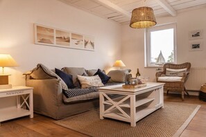A cozy living room with a sofa, a coffee table, and a wicker chair. There are two lamps on side tables, a window with a view, and a wall with framed pictures. - 50111 Fisherman's Loft (Carolinensiel)