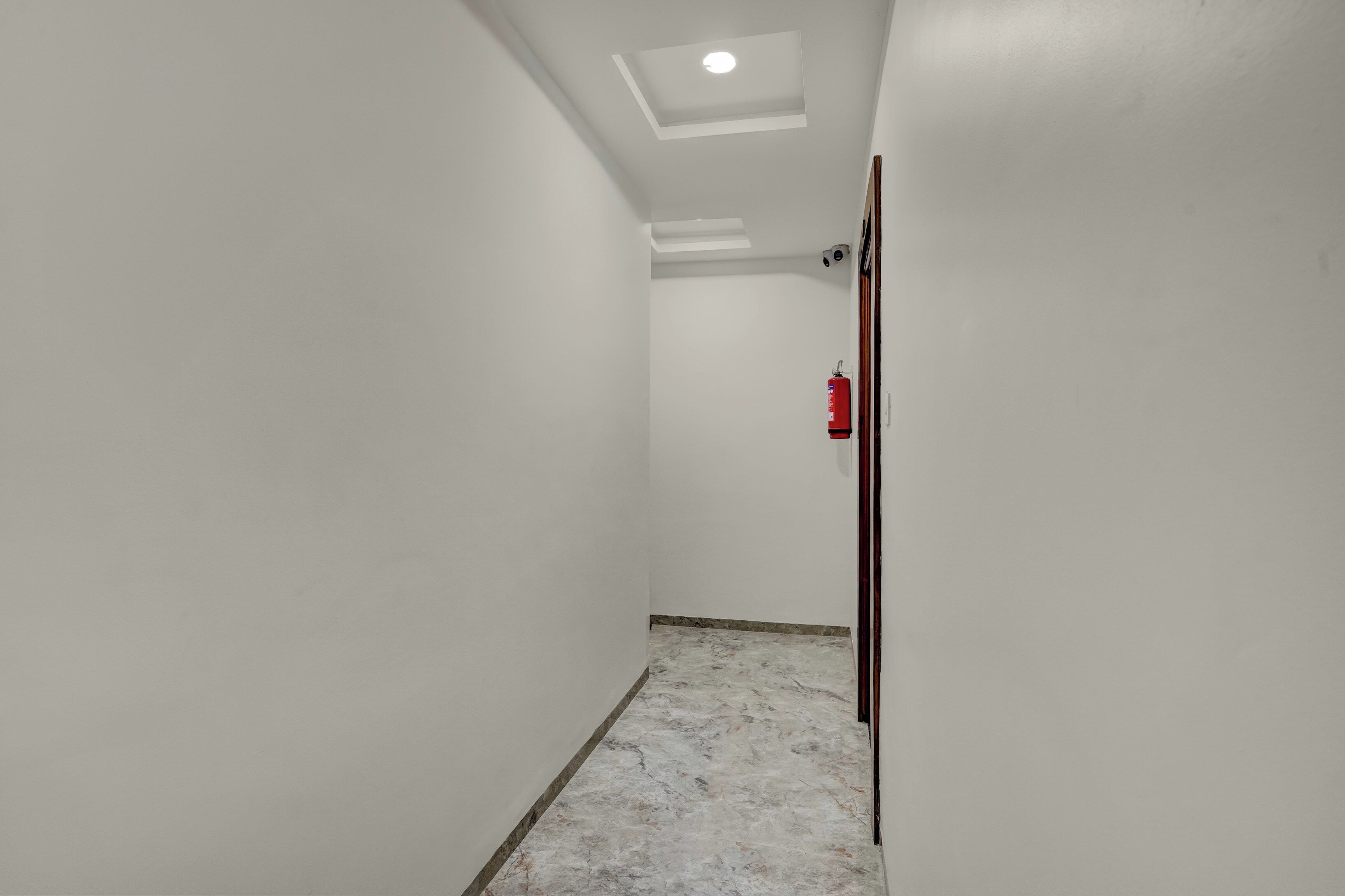 Un pasillo estrecho con paredes blancas, una lámpara en el techo, un extintor y una puerta a la derecha.