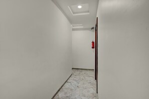 Un couloir étroit aux murs blancs, avec un luminaire au plafond, un extincteur et une porte sur le côté droit.