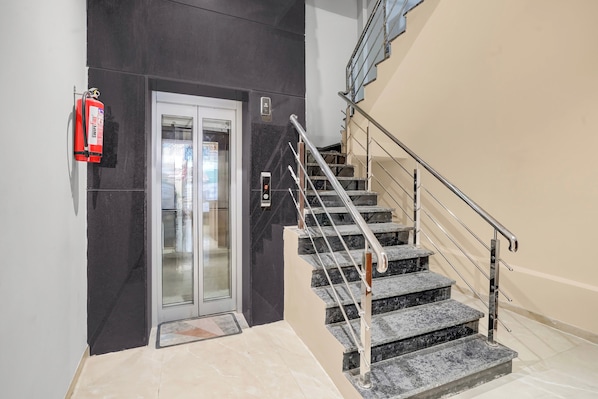 A modern elevator with a glass door, a fire extinguisher mounted on the wall, and a staircase with metal railings.