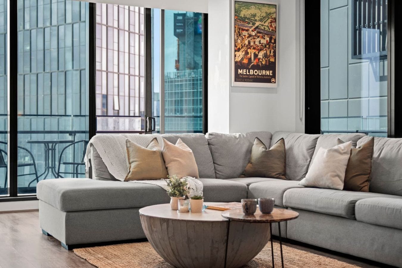 A modern living room with a grey sectional sofa, a round coffee table, and a poster of Melbourne.
