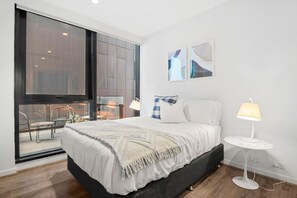 Ein modernes Schlafzimmer mit einem großen Bett, einem kleinen Nachttisch und Blick auf einen Balkon mit Sitzgelegenheiten.