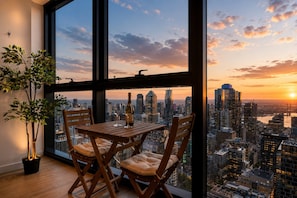 A dining area with a table and chairs, a bottle of wine, and a glass, overlooking a cityscape at sunset. - Sky High 3 Bedroom City Stay (Melbourne)