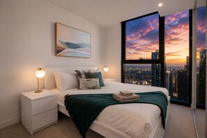 Une chambre moderne avec vue sur la ville, un lit recouvert d’une couverture verte, deux lampes de chevet et un tableau accroché au mur.