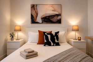 Cama bem arrumada com edredom branco, duas almofadas e um cobertor. Duas mesas de cabeceira com luminárias e uma pequena planta em uma delas. Um quadro pendurado acima da cama.