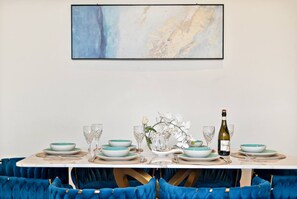 A dining table set with plates, glasses, and a bottle, surrounded by blue upholstered chairs. - Port Melbourne 4 Bedroom Home (Port Melbourne)
