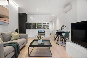 A modern living room with a sofa, coffee table, and TV. A kitchen with white cabinets and a dining area with a table and chairs are visible in the background. - Modern 1 Bedroom City Escape (Melbourne)