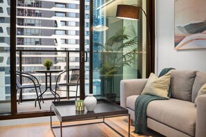 A modern living room with a sofa, a coffee table, and a balcony with a view of a building. - Modern 1 Bedroom City Escape (Melbourne)