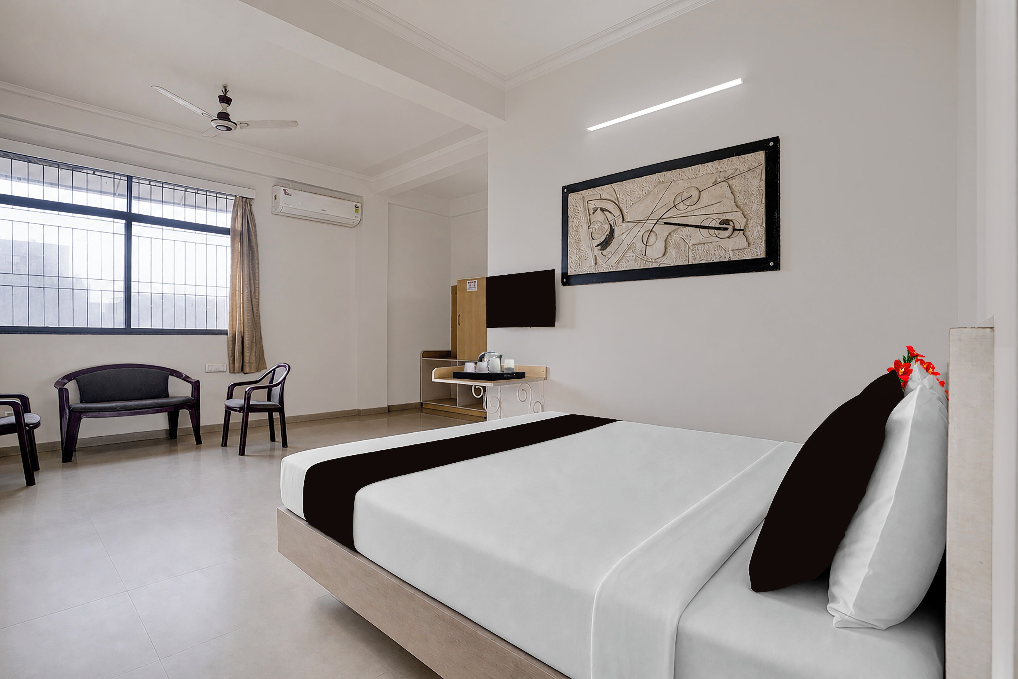 A hotel room with a bed, a desk, a chair, a window with curtains, an air conditioning unit, and a wall-mounted artwork.