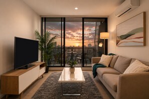 A modern living room with a TV, a coffee table, a sofa, and a balcony with a city view. - 27th Floor Flagstaff 2 Bedroom (Melbourne)