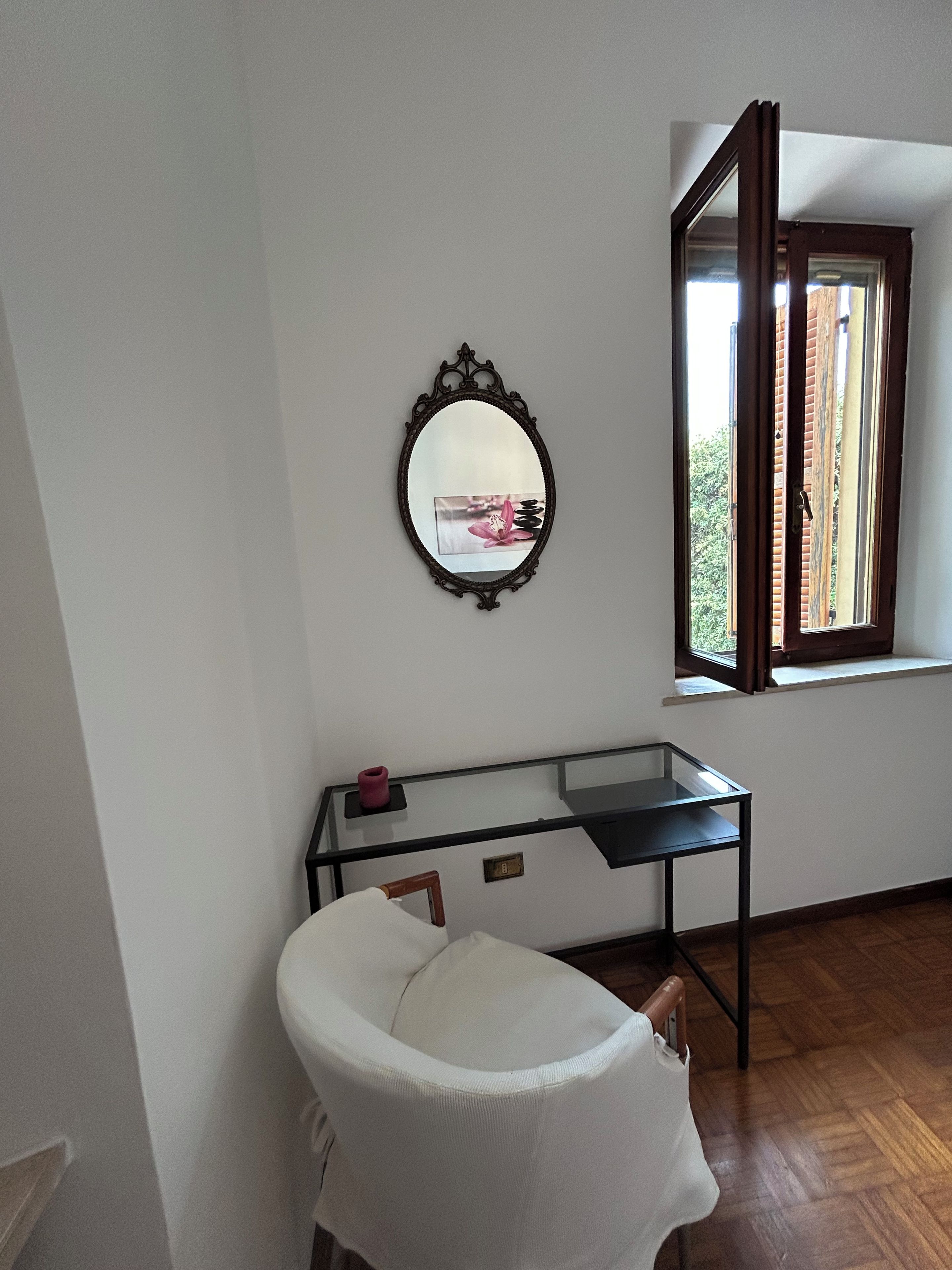 Ein Zimmer mit einem weißen Stuhl, einem Glastisch, einem wandmontierten Spiegel und einem Fenster mit geöffnetem Klappladen.