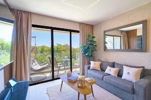 A modern living room with a sofa, coffee table, and a large window with a view of greenery. - Modern 2-bedroom, 2-bathroom condo inside gated Cap Cana. Room for the whole family, or a private bedroom for each couple. Minutes from Juanillo Beach and El Dorado Water Park, 10 min from the airport. (Punta Cana)