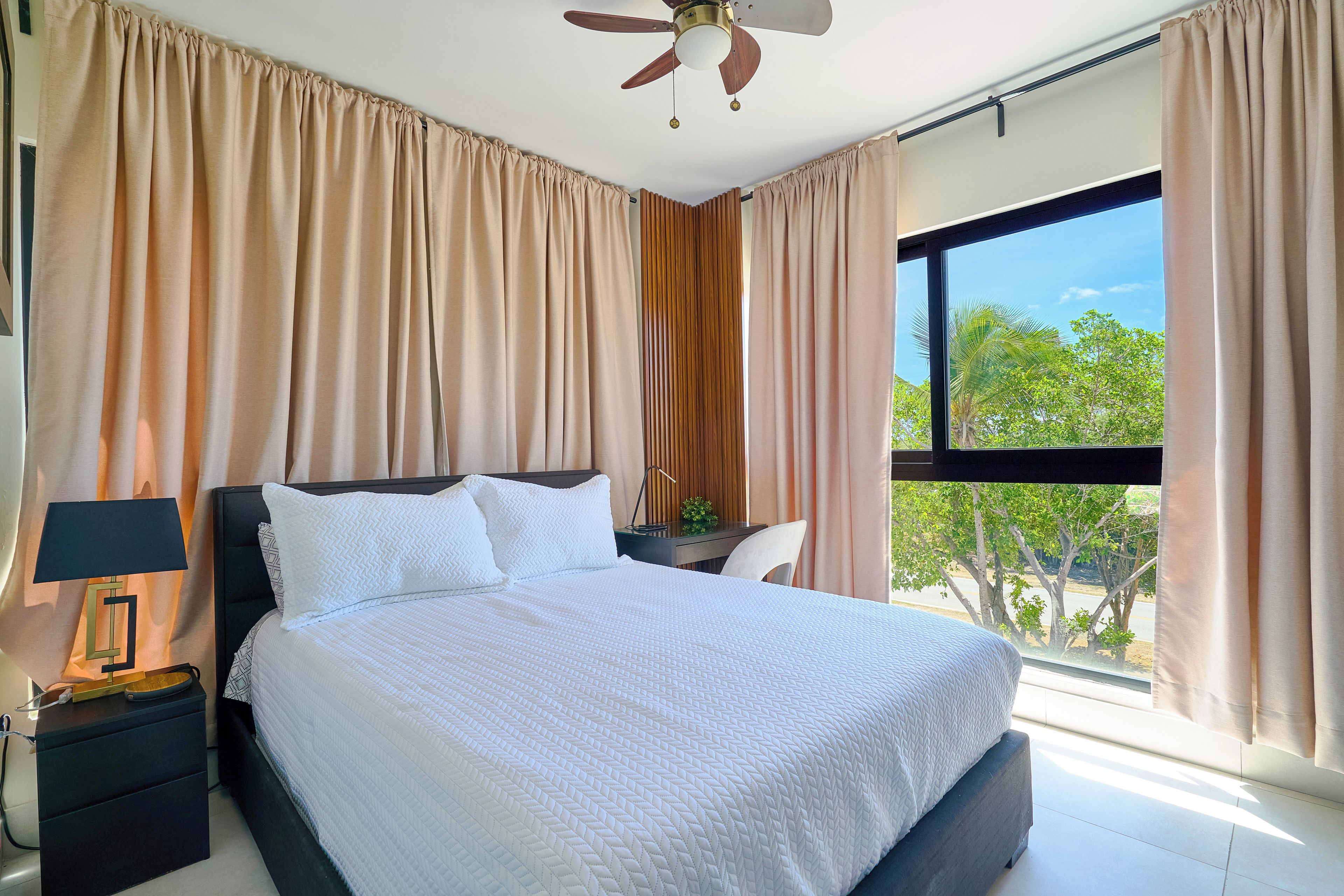 A bedroom with a bed, a nightstand, a lamp, a ceiling fan, and a view of greenery outside.
