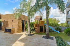 A modern outdoor area with a play structure, a wooden bench, and a small building with a slide. - Modern 2-bedroom, 2-bathroom condo inside gated Cap Cana. Room for the whole family, or a private bedroom for each couple. Minutes from Juanillo Beach and El Dorado Water Park, 10 min from the airport. (Punta Cana)