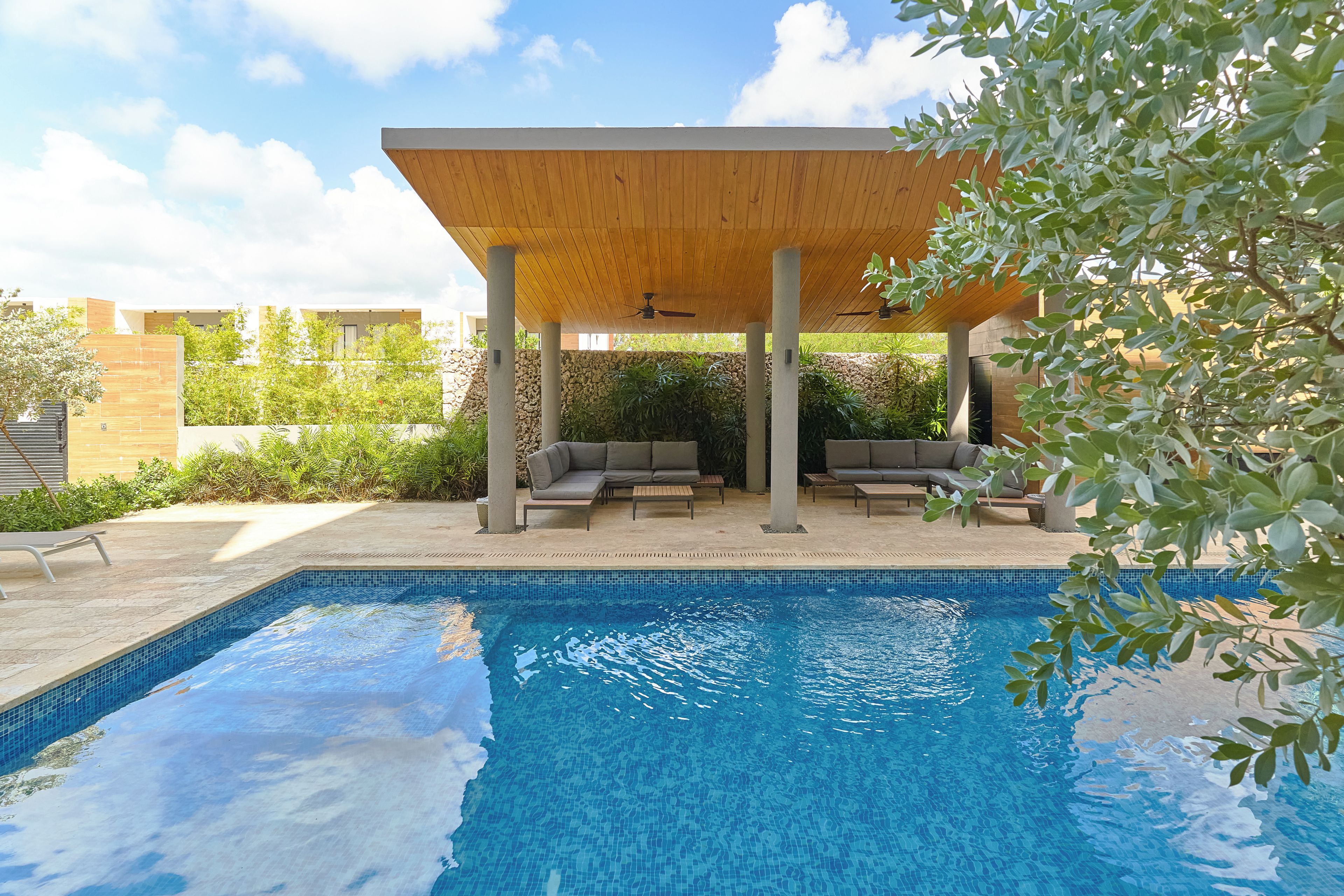 A swimming pool with a wooden pergola, lounge chairs, and a stone wall in the background.