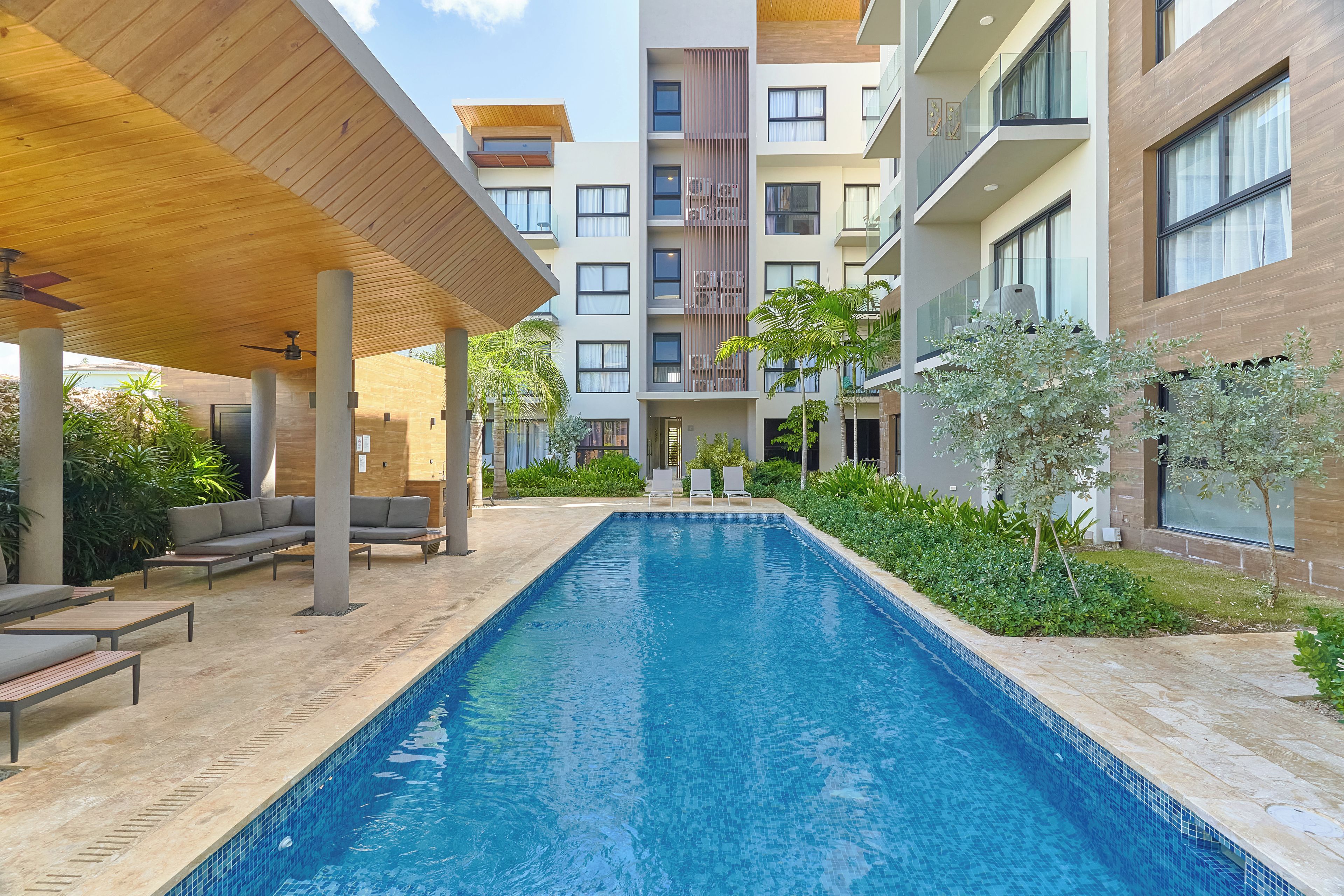 A swimming pool with lounge chairs, a covered area, and a multi-story building in the background.