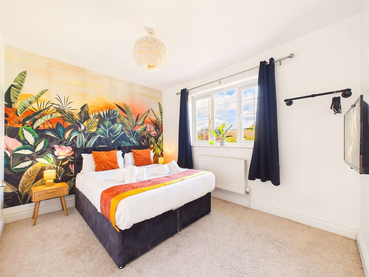 A bedroom with a bed, a window with curtains, a radiator, and a wall with a tropical mural.