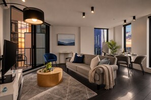 A modern living room with a sofa, coffee table, and TV. There is a dining area with a table set for two. Large windows offer views of the city. - Stadium Side 2 Bedroom Retreat (Docklands)