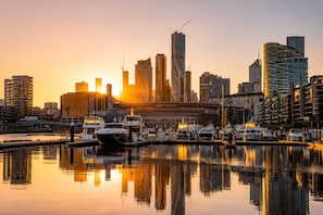A marina with boats docked, a city skyline with modern buildings, and a sunset in the background. - Stadium Side 2 Bedroom Retreat (Docklands)