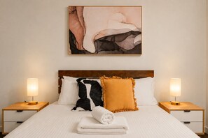 A neatly made bed with a white comforter, two pillows, a rolled white towel, and two bedside lamps on wooden nightstands. - Stadium Side 2 Bedroom Retreat (Docklands)