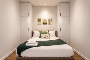 Un dormitorio con cama, paredes blancas y obras de arte sobre la cama.