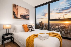 A hotel room with a large bed, a nightstand, a lamp, a city view, and a painting on the wall. - 70th Floor City View Apartment (Melbourne)