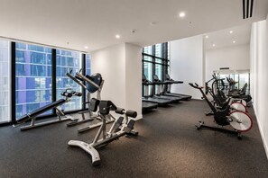 A gym with various exercise machines, large windows, and a carpeted floor. - 70th Floor City View Apartment (Melbourne)