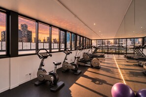 A gym with stationary bikes, treadmills, and exercise balls, with a city skyline view through large windows. - Cozy Docklands 1 Bedroom (Docklands)