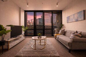 A modern living room with a flat-screen TV, a sofa, a coffee table, and a large window with a city view. - Cozy Docklands 1 Bedroom (Docklands)